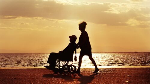 How to Travel with Someone in a Wheelchair - Woman Pushing Man in Wheelchair on a Beach at Sunset