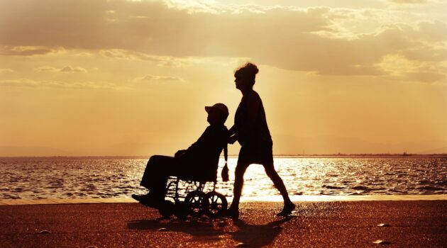 Homepage 2 How to Travel with Someone in a Wheelchair - Woman Pushing Man in Wheelchair on a Beach at Sunset